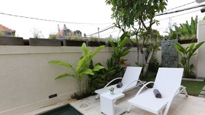 A poolside area with two white lounge chairs, a small table with a vase, and a stone wall with greenery. - Green Studio Sanur (Bali)