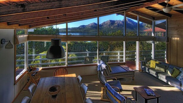 A spacious room with a large window offering a view of mountains and a lake. There is a wooden table, a sofa, and a bench. - Freycinet Beachfront Retreat - Freycinet Holiday Houses (Coles Bay)