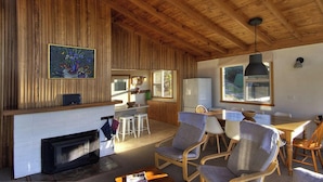 A rustic interior with a wooden ceiling, a fireplace, a dining area with chairs, and a kitchen with a refrigerator. - Freycinet Beachfront Retreat - Freycinet Holiday Houses (Coles Bay)