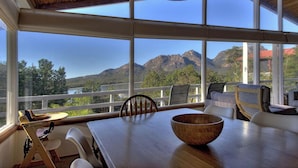 A dining area with a wooden table and chairs, a wooden bowl, and a view of mountains and greenery through large windows. - Freycinet Beachfront Retreat - Freycinet Holiday Houses (Coles Bay)