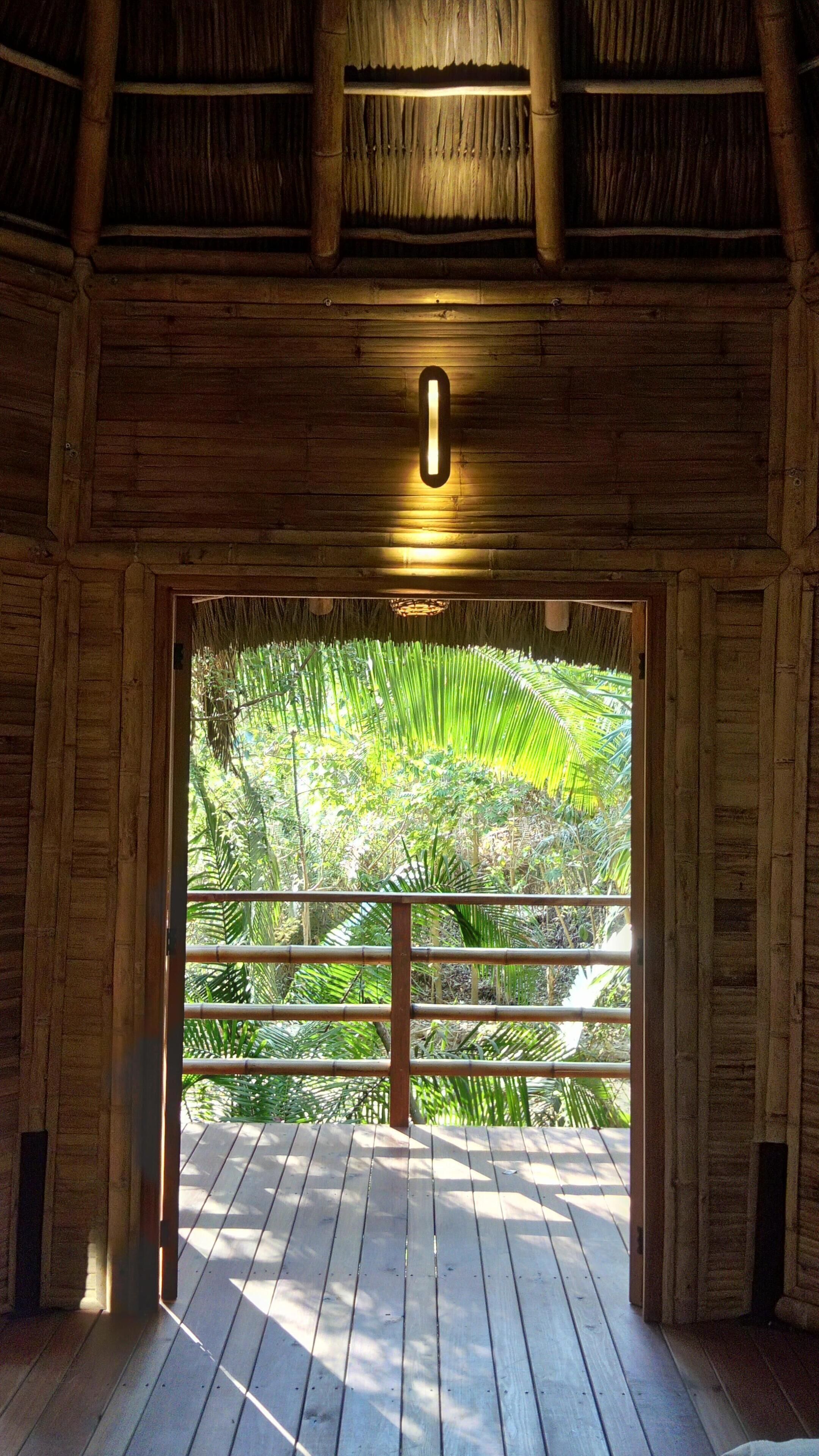 Une structure en bois surmontée d’un toit de chaume, dotée d’un luminaire central et offrant une vue sur un jardin luxuriant grâce à une porte en bois.