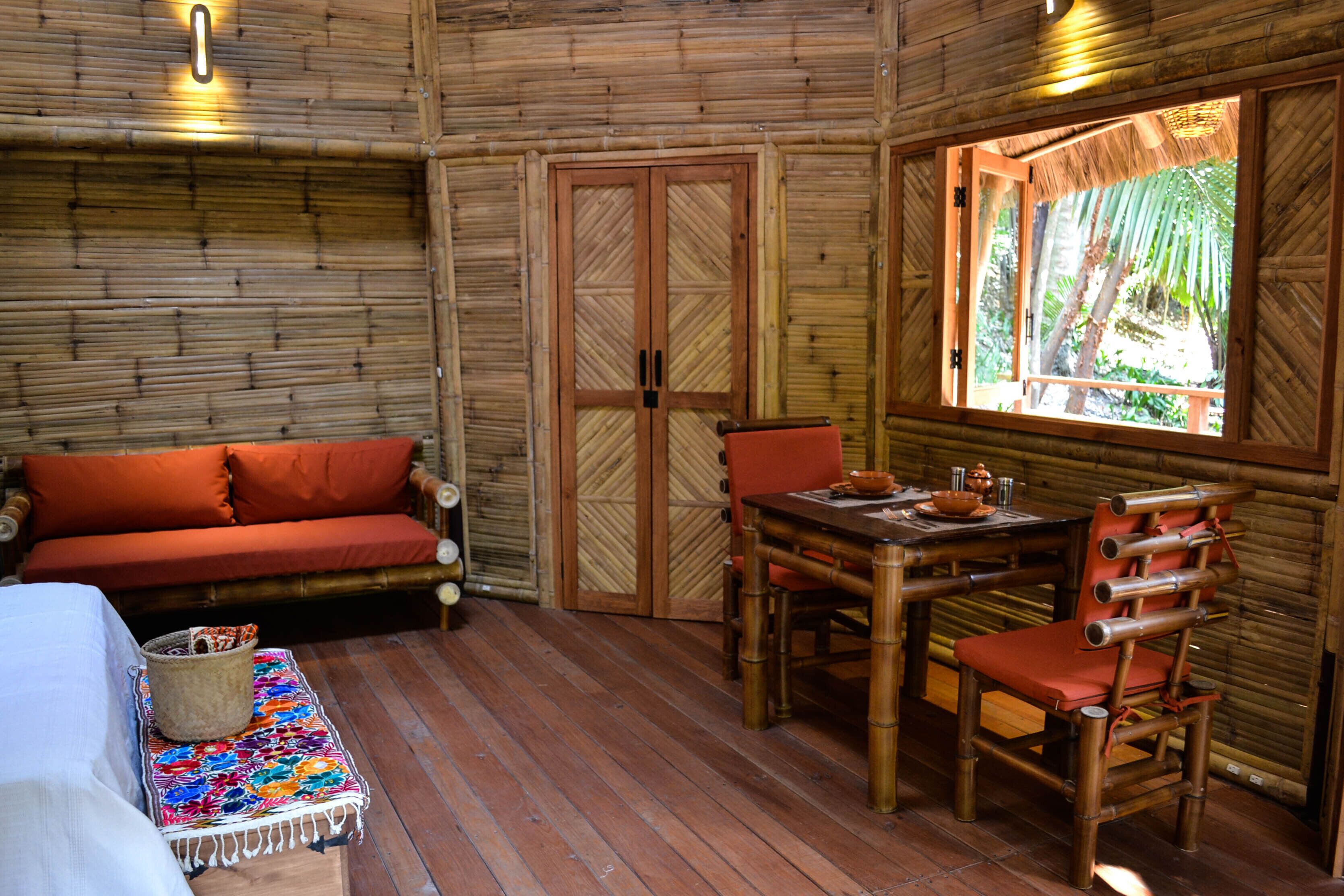 A room with a wooden floor, a sofa with orange cushions, a dining table with chairs, and a window with a view of greenery.