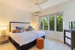 A double bed with a headboard, two bedside lamps, a ceiling fan, a window with a view of greenery, and a TV stand.