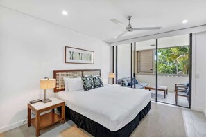Ein Schlafzimmer mit einem Bett, einem Nachttisch, einem Deckenventilator, einem Fenster mit Blick auf Grünflächen und einer Glastür, die zu einem Außenbereich führt.