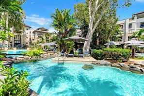 Ein Poolbereich mit Liegestühlen, Sonnenschirmen und einem klaren, blauen Pool, umgeben von Bäumen und Gebäuden.