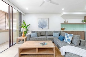 A modern living room with a grey sectional sofa, a wooden coffee table, and a large window with blinds.