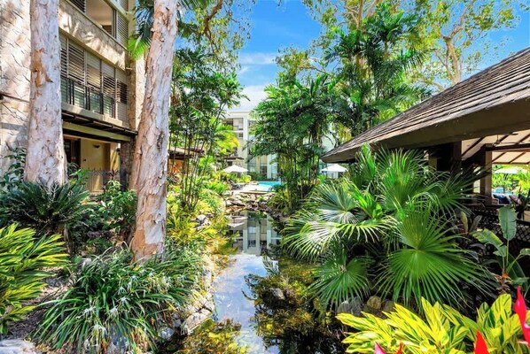 A lush garden with a water feature, a pool, and a building with balconies.