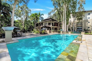 A swimming pool with lounge chairs and umbrellas, surrounded by trees and a building with balconies.