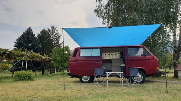 A red van with a blue awning, a small table, and a chair set up in a grassy area with trees in the background.