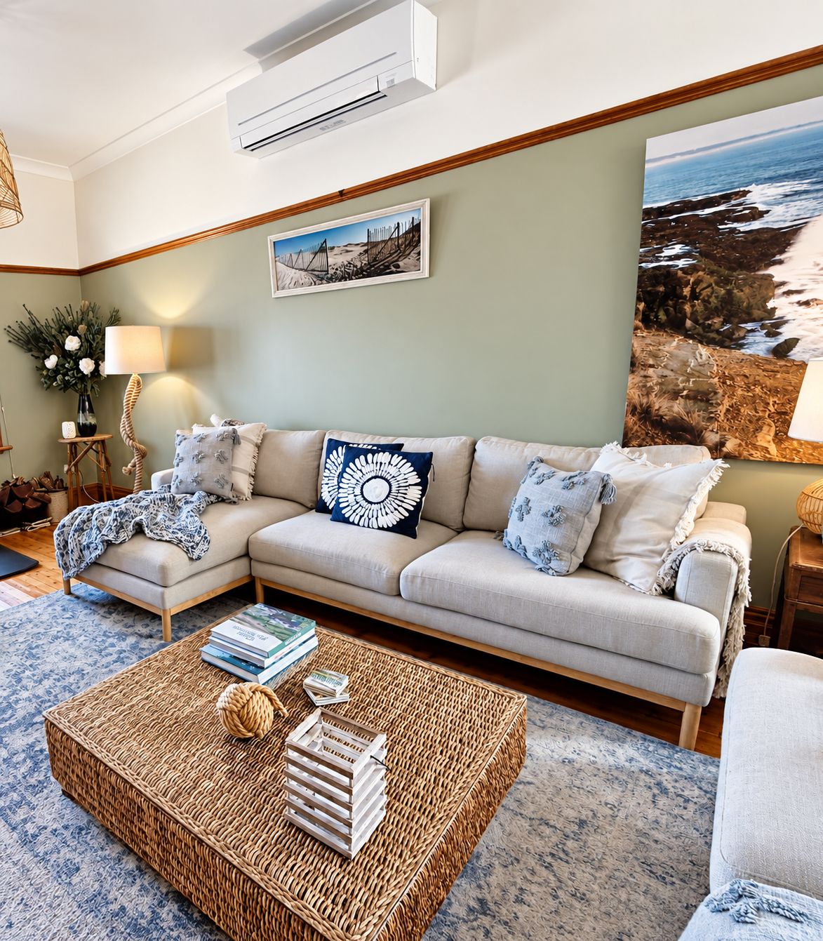 A living room with a beige sectional sofa, a woven coffee table with books, a wall-mounted air conditioner, and a large painting of a coastal scene.