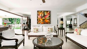 Un salon spacieux avec un canapé blanc, deux fauteuils, une table basse en verre et un ventilateur de plafond. Un grand tableau représentant des fruits est accroché au mur, et un escalier est visible au fond.