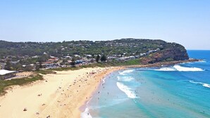 Una playa costera con arena, aguas azules cristalinas y una colina con edificios.
