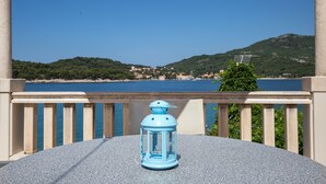 Un balcon avec une table et une lanterne bleue, donnant sur un paysage côtier avec des collines et une étendue d’eau.
