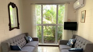 A room with two grey sofas, a large window with a view of greenery, a wall-mounted TV, and an air conditioning unit.