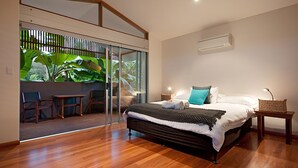 A modern bedroom with a large bed, a glass door leading to a balcony with plants, a small table, and a nightstand with a lamp.