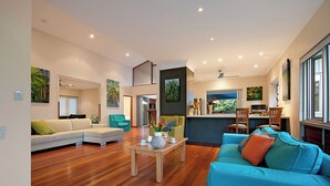A spacious living area with a wooden floor, a blue sofa, a white sofa, and a kitchen area with a dining table and chairs.