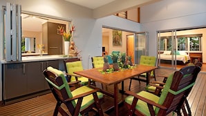 A modern dining area with a wooden table and chairs, a kitchen with sliding doors, and a view of a bedroom with a bed and a window.