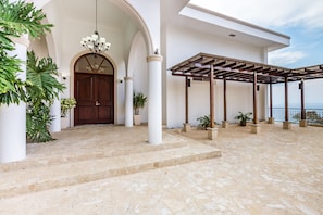 A stone-floored entrance with a wooden door, white columns, and a pergola with a view of the sea.