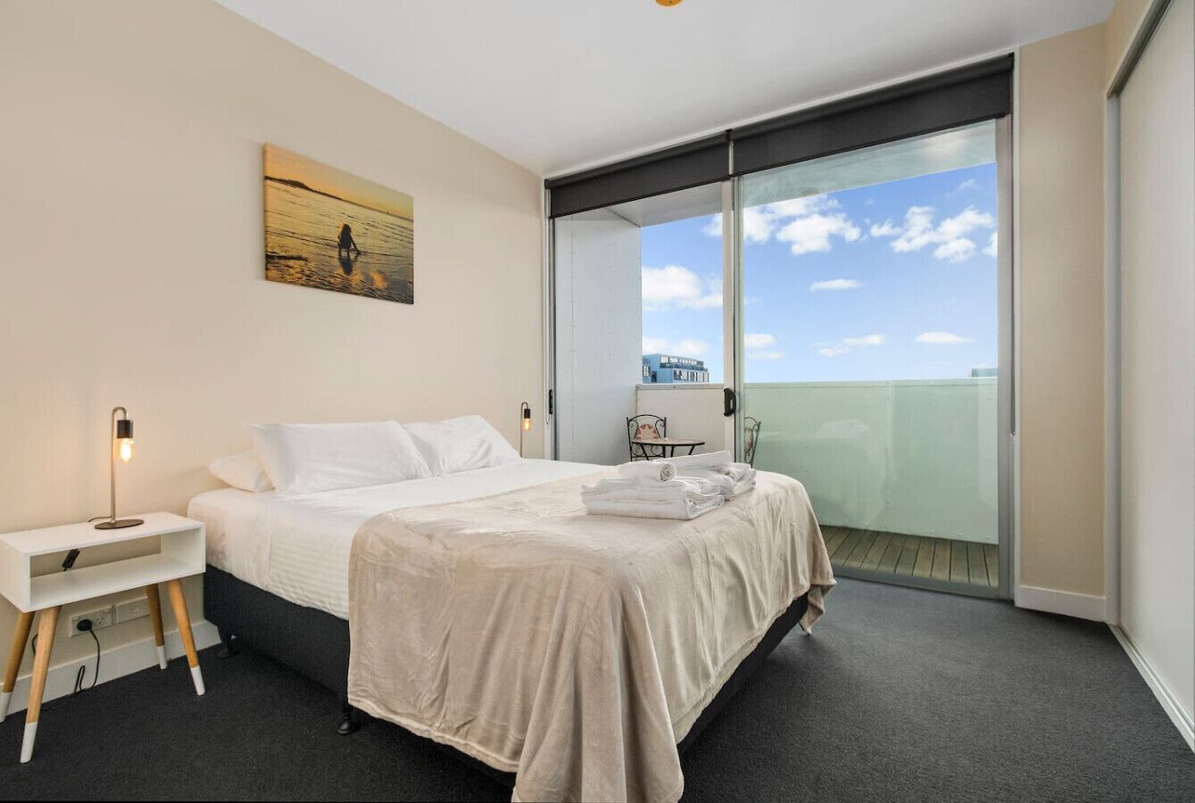 A hotel room with a bed, a bedside table with a lamp, a painting on the wall, and a view of the sky through a large window.