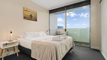 A hotel room with a bed, a bedside table with a lamp, a painting on the wall, and a view of the sky through a large window.