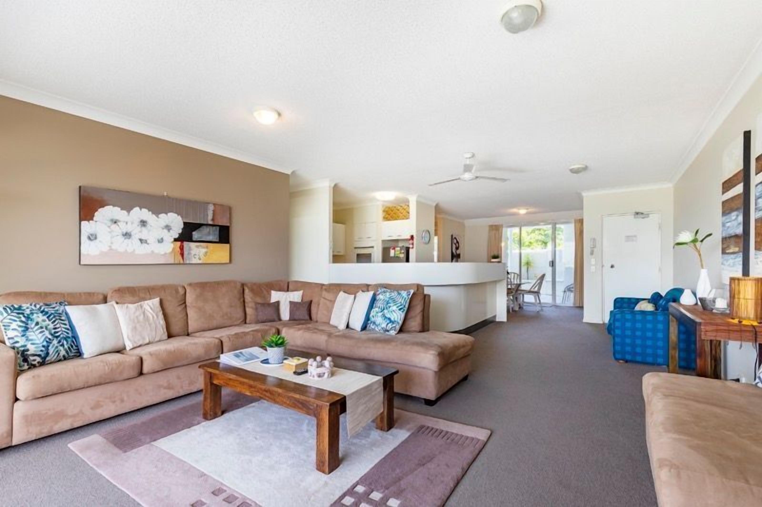 A living room with a large beige sectional sofa, a wooden coffee table, and a blue armchair. There is a kitchen in the background with white cabinets and a dining area.