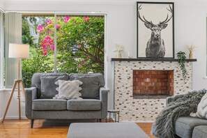 A living room with a grey sofa, a fireplace, and a large window with pink flowers. - Salt & Sand - Flynn's Beach (Port Macquarie)