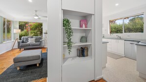 Un intérieur moderne avec un canapé gris, un petit pouf, une bibliothèque remplie de livres et d’objets décoratifs, une cuisine avec des meubles blancs et une vue sur la verdure à l’extérieur.
