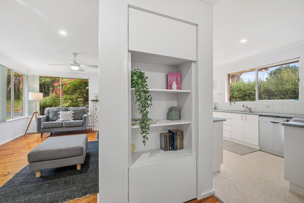 A modern interior with a grey sofa, a small ottoman, a bookshelf with books and decorative items, a kitchen with white cabinets, and a view of greenery outside. - Salt & Sand - Flynn's Beach (Port Macquarie)