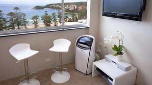 A room with a large window offering a view of the sea and buildings, a white TV on the wall, and two white bar stools.