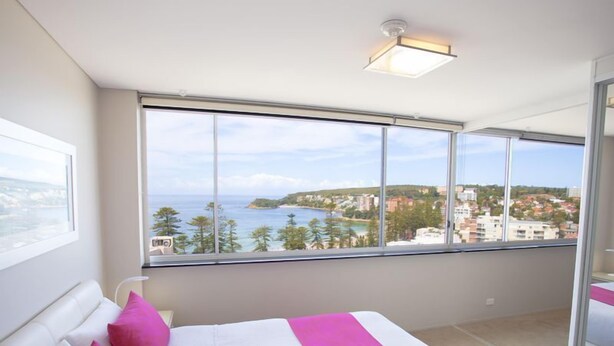 A bedroom with a large window offering a view of the sea and buildings.