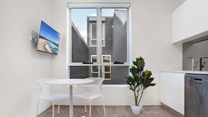 A modern kitchen with a built-in dishwasher, a dining area with a white table and chairs, and a large window offering a view of the outside. - BONDI BEACH STUDIOS: VII (Bondi)
