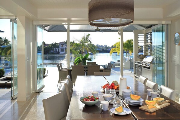 A dining area with a view of a pool and palm trees, featuring a table set with fruits and a grill. - The Peninsula Secret (Noosaville)