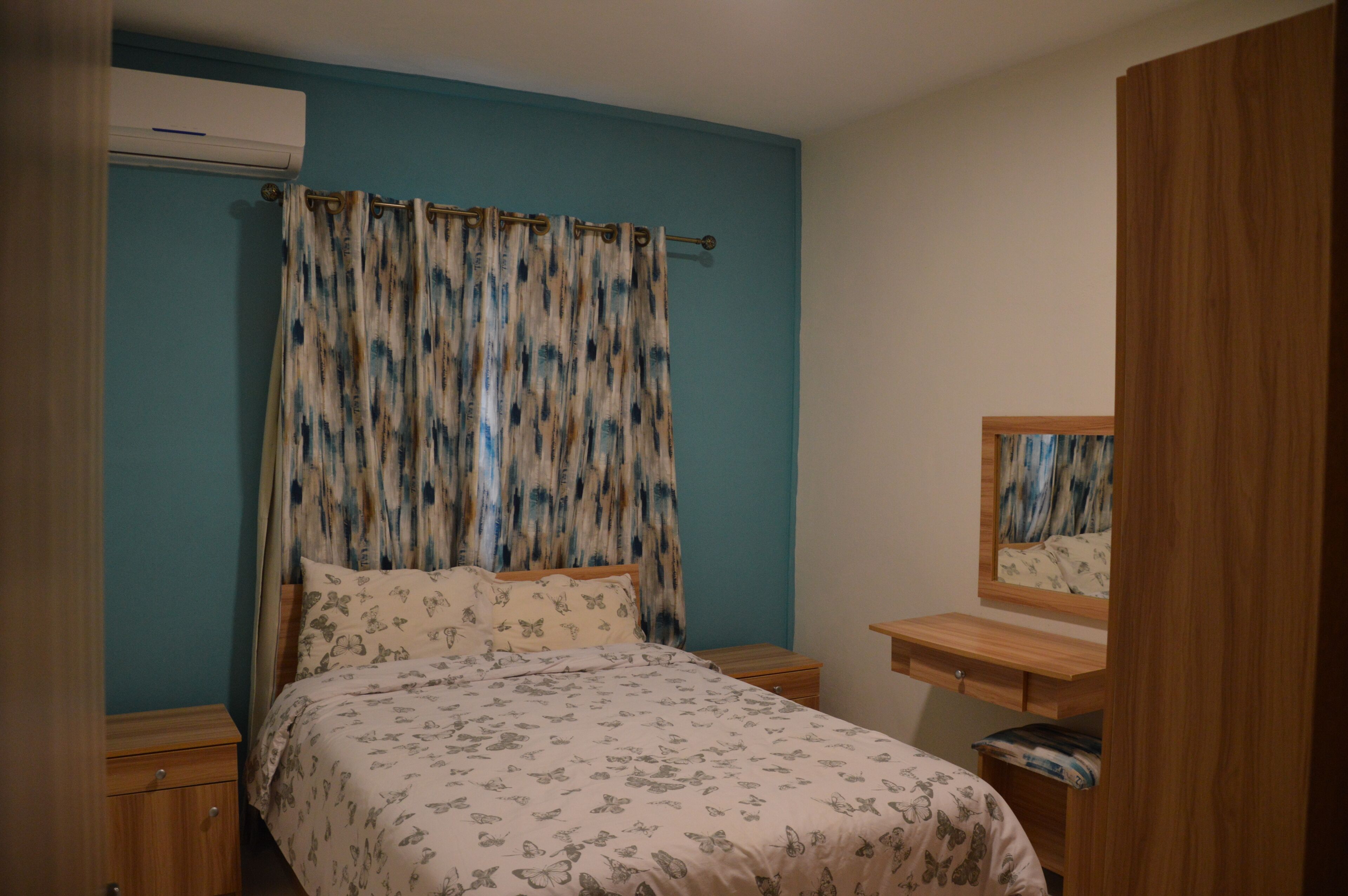 Una camera da letto con un letto, un comodino, una cassettiera in legno, uno specchio e una finestra con tende.