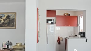 A compact kitchen with a refrigerator, microwave, and red cabinets. There is a small countertop with a toaster and a kettle. A painting and a lamp are on a table to the left.