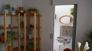 A dining area with a table set for two, a wooden plant shelf, and a view into a bathroom with a sink and mirror.