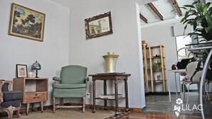 A room with a green armchair, a wooden side table with a brass vase, a framed mirror, and a painting on the wall.