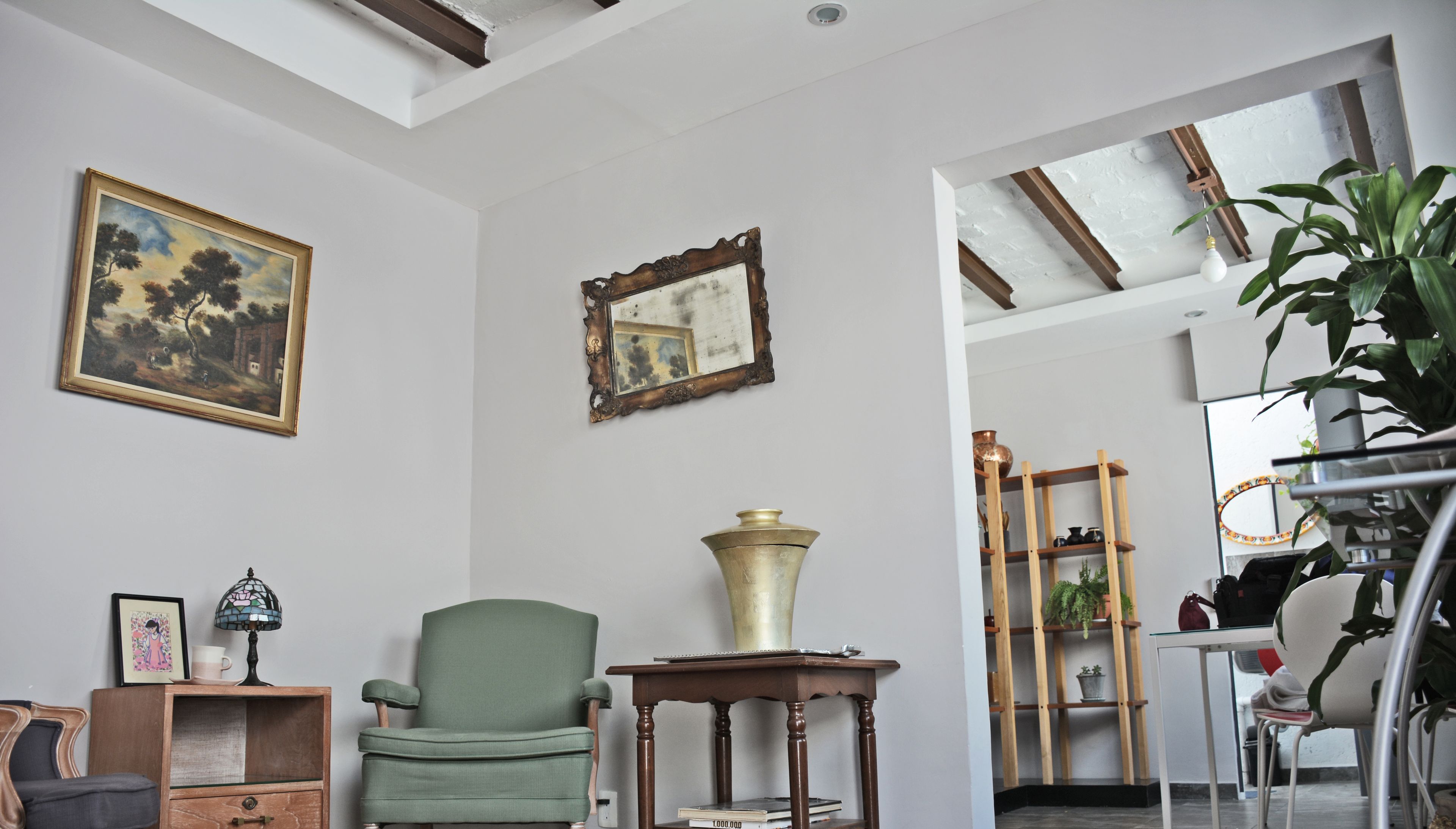 A room with a green armchair, a wooden side table with a brass vase, a framed mirror, and a painting on the wall.