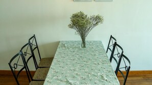 A dining area with a table covered in a floral tablecloth, surrounded by four chairs, a vase with flowers, and a hanging light fixture.