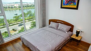 A bedroom with a bed, a nightstand, a lamp, and a view of a river and buildings through large windows.
