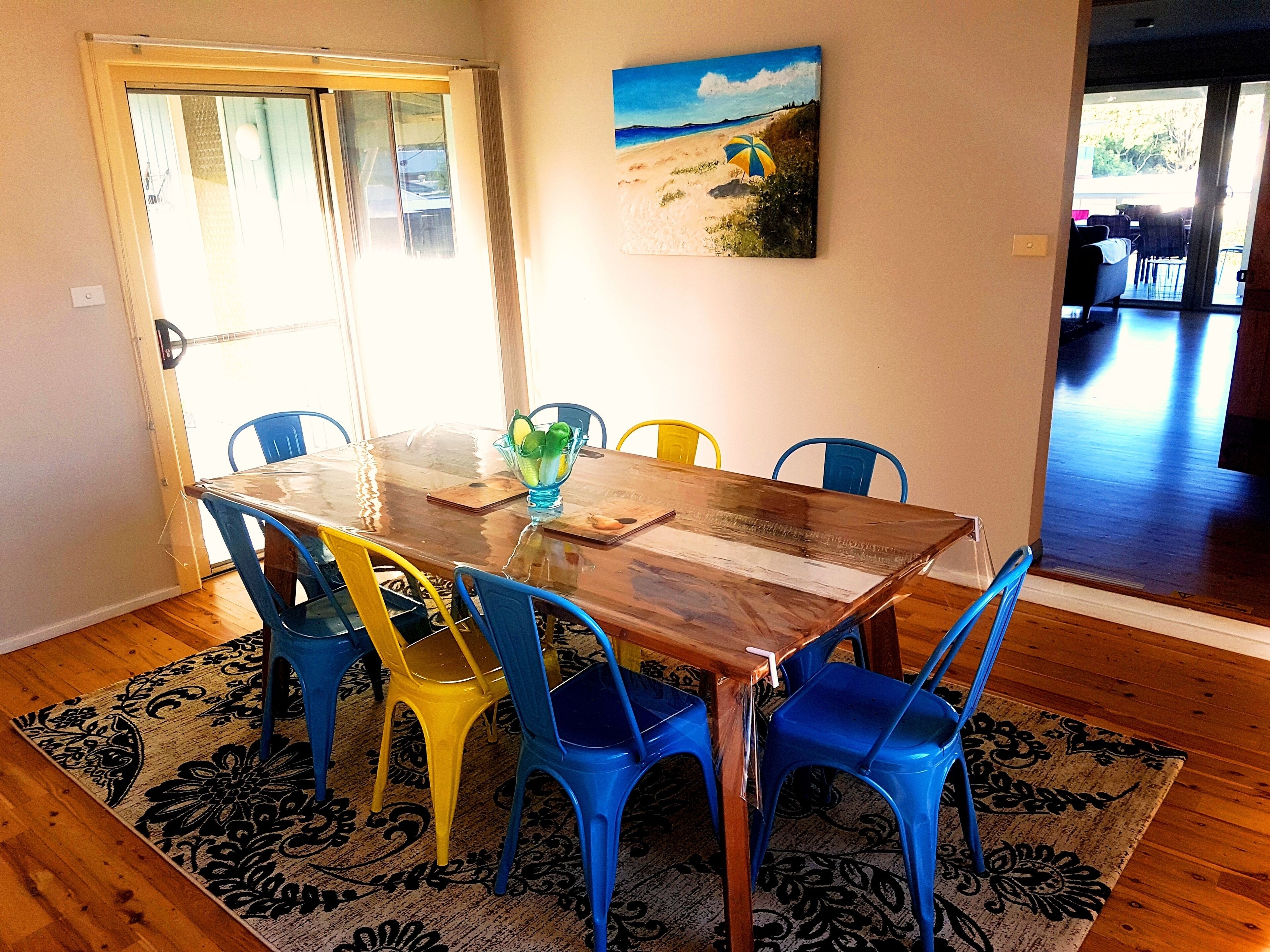 A dining area with a glass-top table, colorful chairs, a fruit bowl, and a beach-themed painting on the wall.