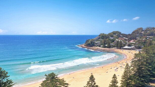 A coastal beach with clear blue waters, sandy shores, and a backdrop of residential buildings and trees.