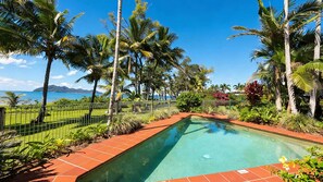 A rectangular pool with a tiled edge, surrounded by a variety of tropical plants and palm trees, with a clear view of the ocean and distant mountains.