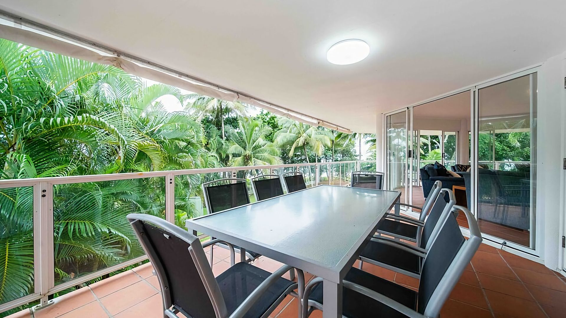 A balcony with a dining table and chairs, surrounded by glass doors and a view of lush greenery.
