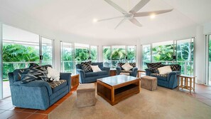 A spacious living area with a ceiling fan, large windows, and a view of lush greenery.