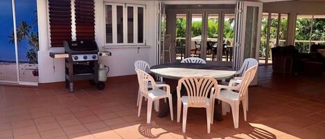 Un patio al aire libre con una mesa redonda y sillas, una parrilla y vista a la playa a través de puertas corredizas de vidrio.