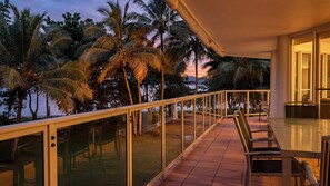 A balcony with a glass railing, overlooking palm trees and a sunset.