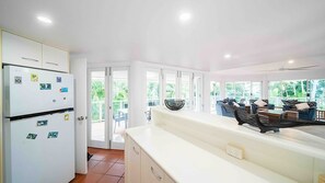 A modern kitchen with a white refrigerator, a dining area with a sofa and a coffee table, and a view of the outdoors through large windows.