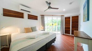 A hotel room with a bed, a ceiling fan, an air conditioning unit, a nightstand, a lamp, a window with curtains, and a view of greenery outside.