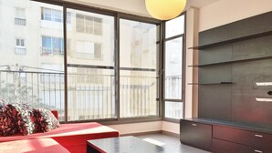 Un salon moderne avec un canapé rouge, une table basse noire et une grande fenêtre donnant sur les bâtiments voisins.