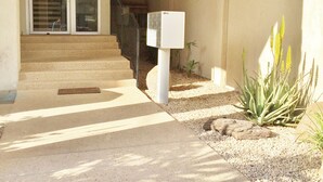 Une entrée d’habitation avec un chemin en béton menant à une porte, une boîte aux lettres et un petit espace jardin agrémenté de plantes.